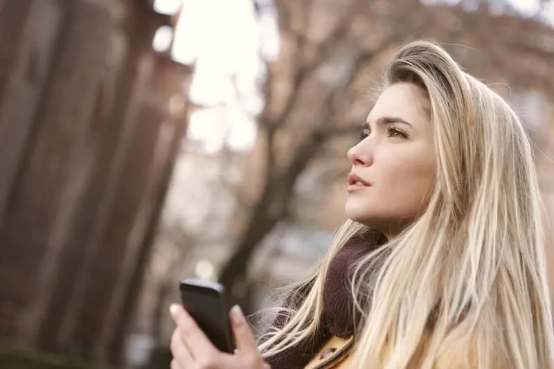 pensive woman standing outdoors holding a smartphone