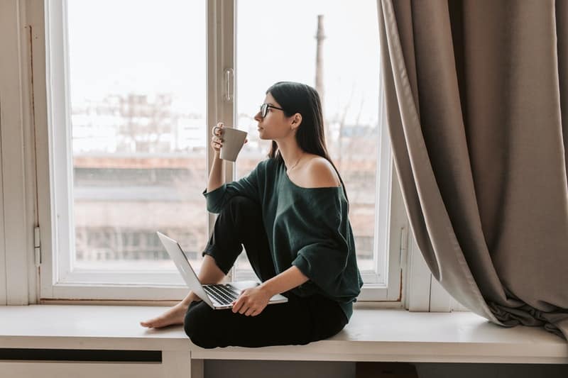 pensive woman with laptop and hot coffee sitting on windowsill