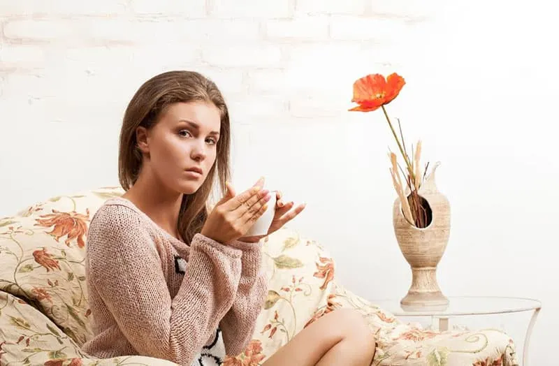 sad woman holding a cup relaxing at couch at home near flower vase
