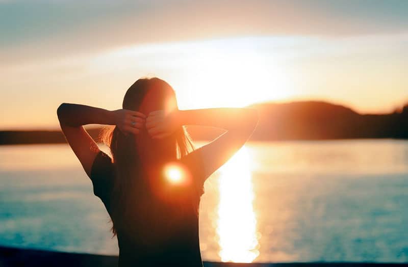 silueta de mujer frente a la puesta de sol y el mar con las manos en la cabeza