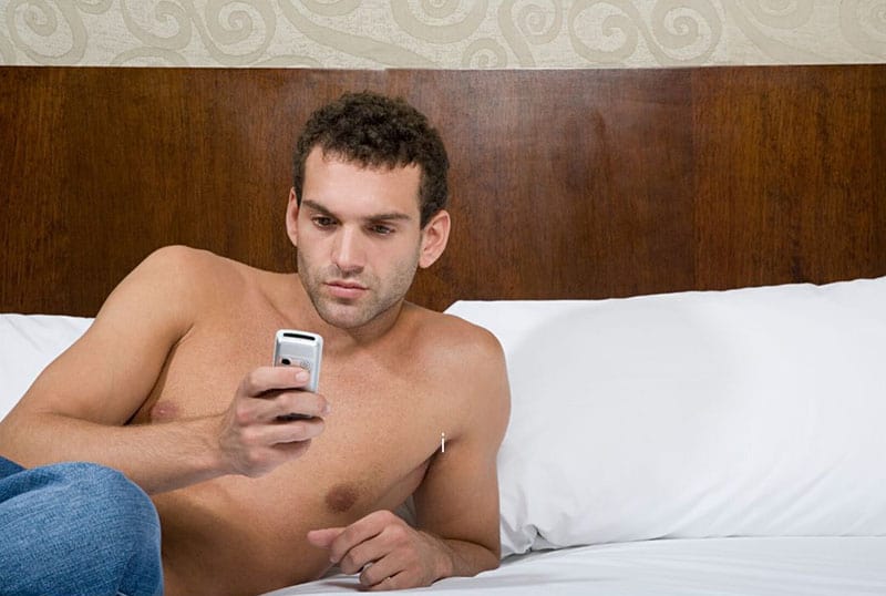 topless man texting on bed with white linen