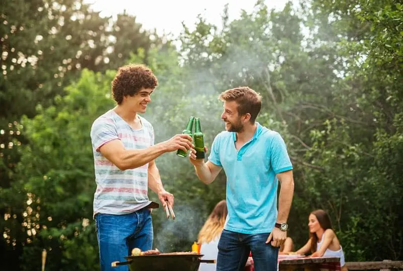 two guys with bro code clinking bottles with some friends at their back during a barbecue party