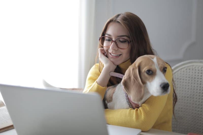 woman embracing her dog while while sitting and checking on her laptop and smiling inside home
