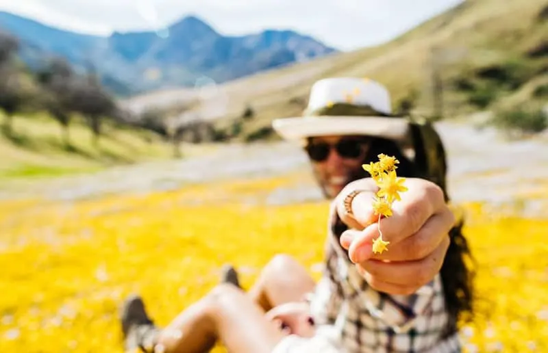 woman holding yellow flower sitting on the ground full of flowers near the mountains and hills
