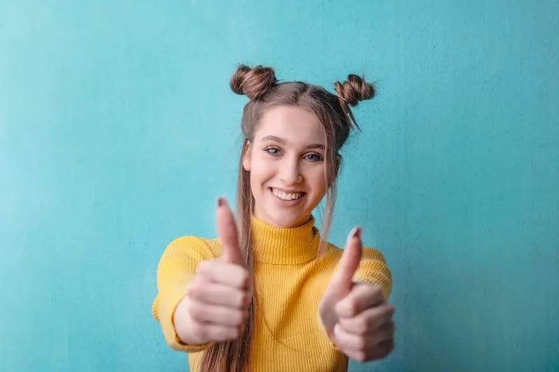 woman in yellow turtleneck sweater smiling and giving two thumbs up standing against green wall