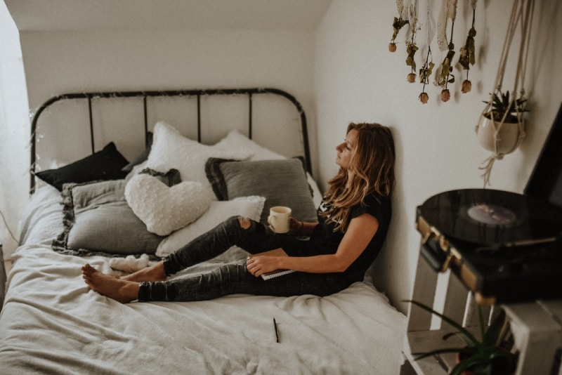 mulher com uma caneca encostada à parede enquanto está sentada na cama