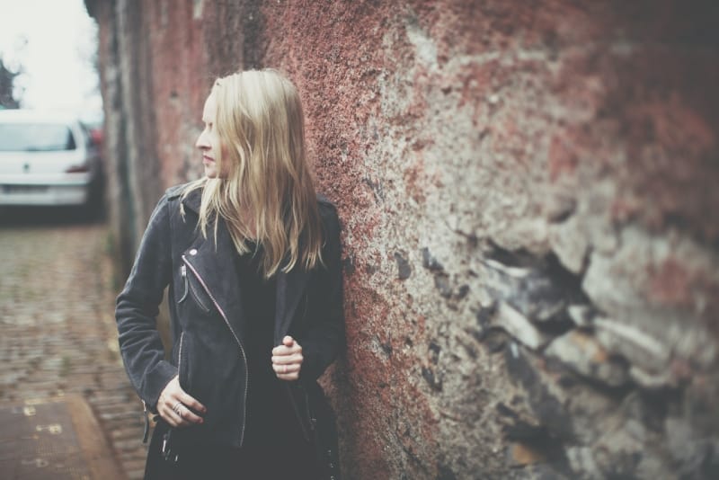 blonde woman in black jacket leaning on brown wall