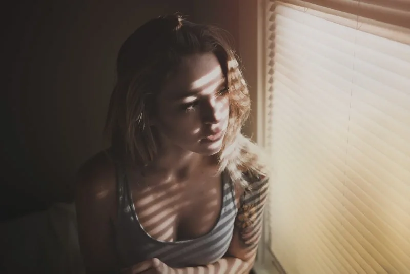woman in gray top looking through window blinds