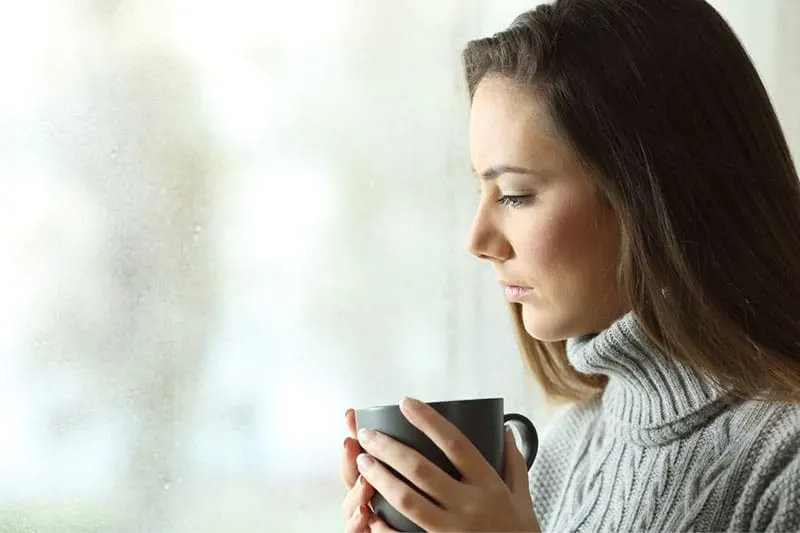 woman near the glass window drinking a cup of coffee while thinking