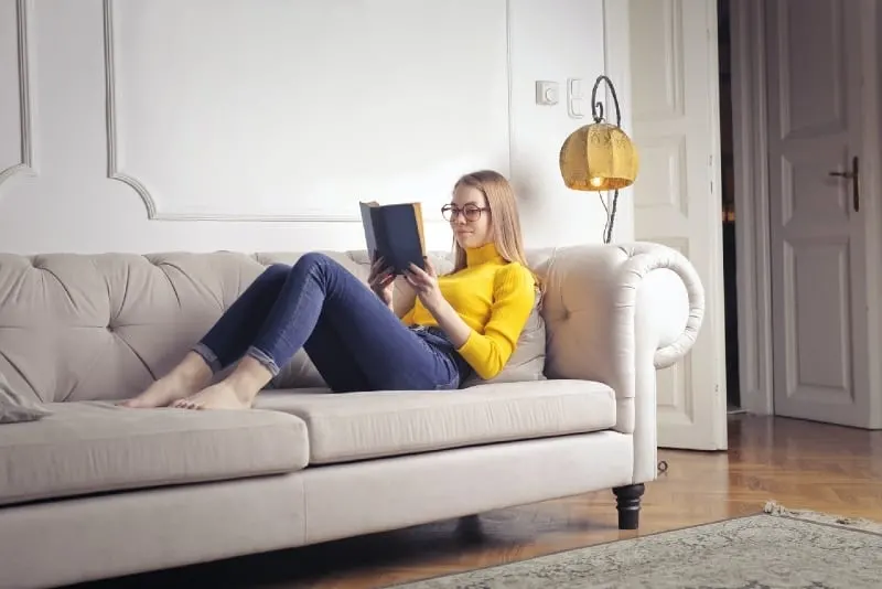 woman with eyeglasses reading book while sitting on sofa
