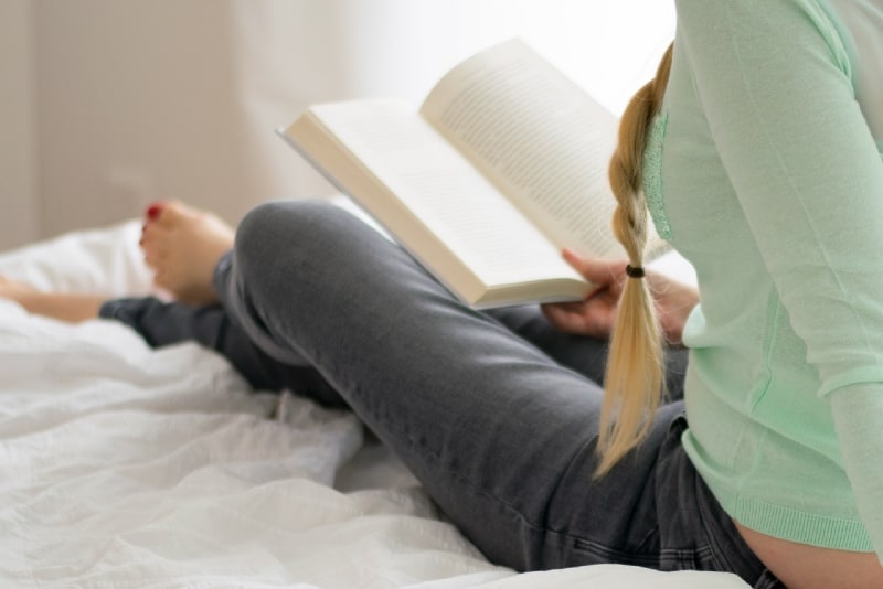 woman reading book while sitting on bed