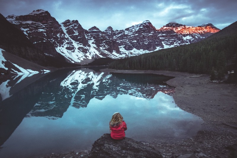 woman in red jacket sitting near lake looking at mountain