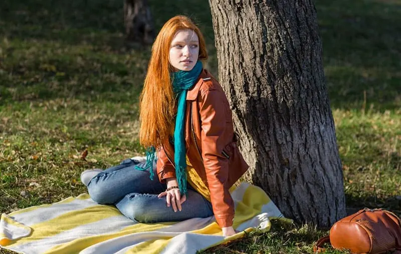 woman sitting on picnic mat under a tree looking far and wearing brown jacket