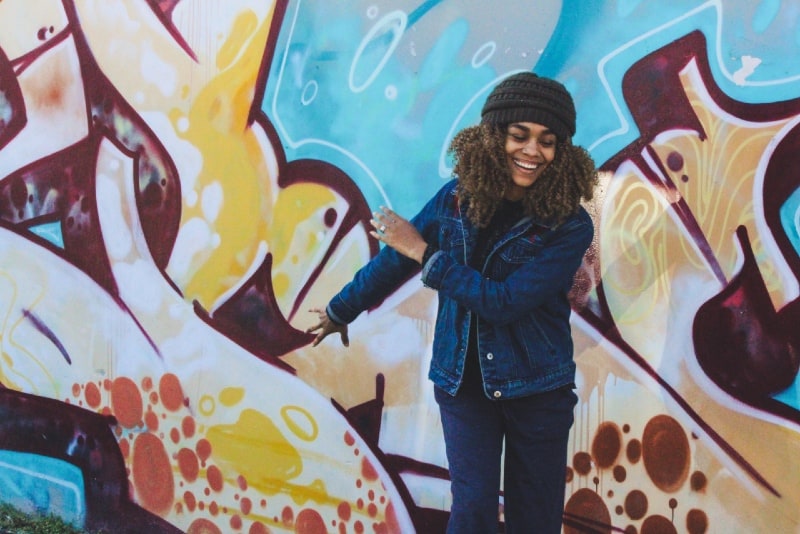 smiling woman in denim jacket standing near graffiti wall