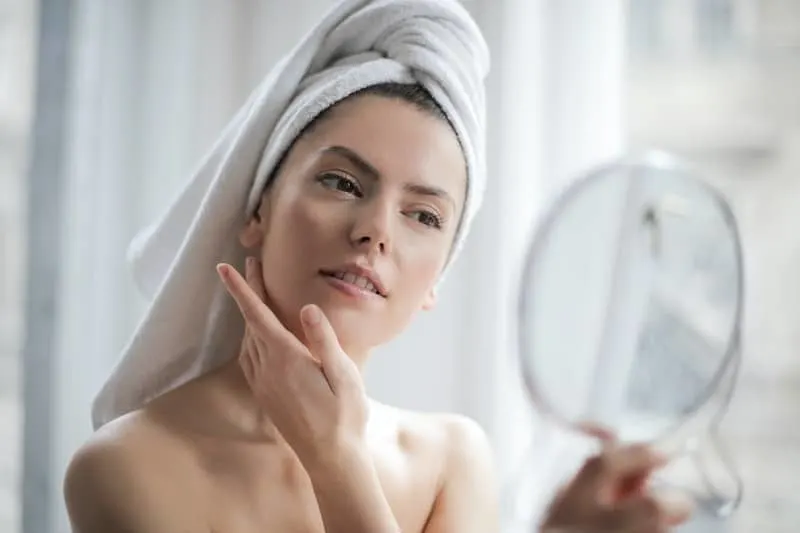 woman with towel on head touching face and facing a small mirror