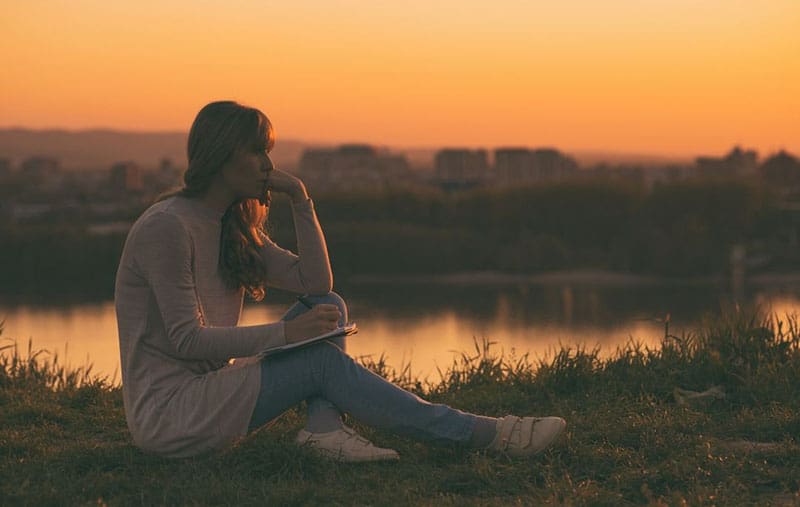 woman writing on notebook sitting on the ground near a body of water during sunset