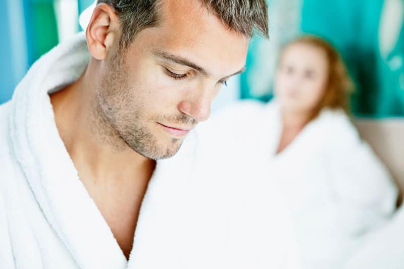 young man in white robe sad with a woman behind him in bed