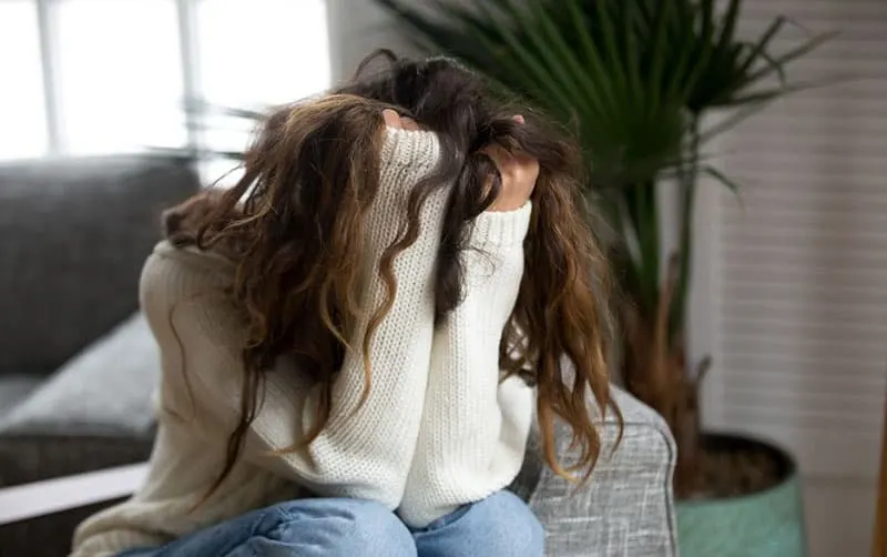 young woman suffering from pain grasping her hair sitting inside living room wearing sweater