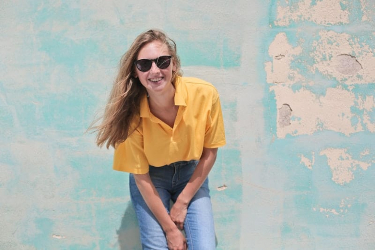 beautiful happy woman wearing yellow top and sunglasses standing near a green wall