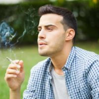 homem a fumar um cigarro ao ar livre com um top axadrezado azul