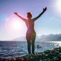mujer levantando las manos de pie en la orilla del mar frente a la salida del sol brillante