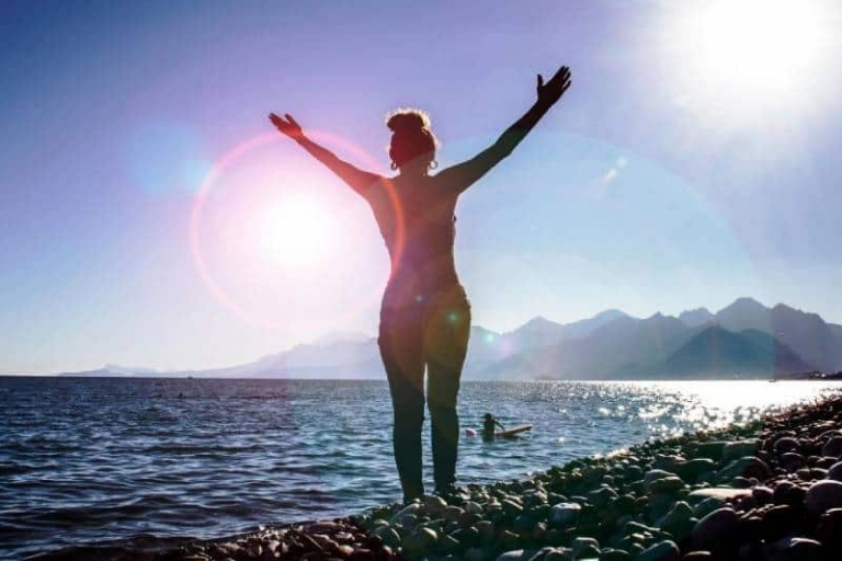 woman raising hands standing in the seashore facing the bright sunrise