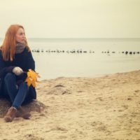 pensive woman sitting on the sand on the beach suring autumn holding dried leaves