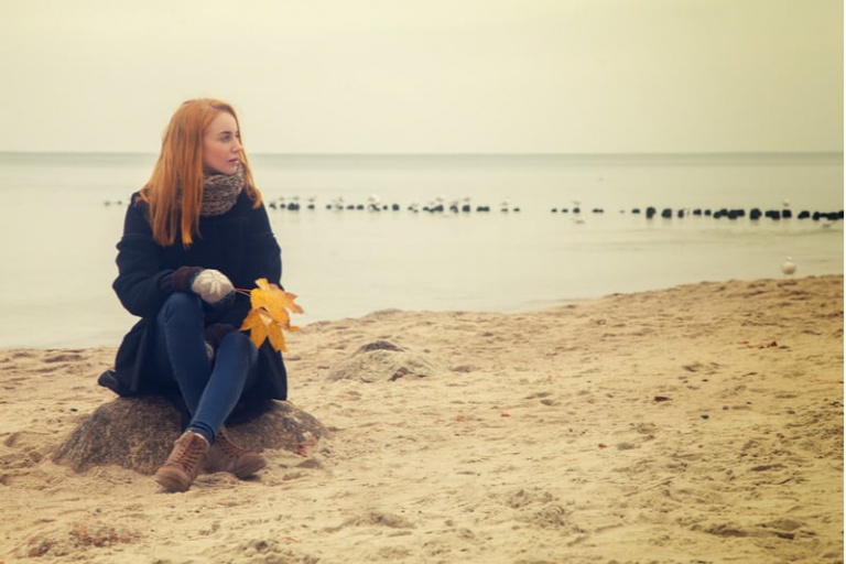 mujer pensativa sentada en la arena de la playa a la entrada del otoño sujetando hojas secas