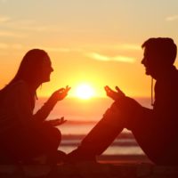 vista lateral de dos amigos de cuerpo entero hablando durante la puesta de sol cerca de una masa de agua