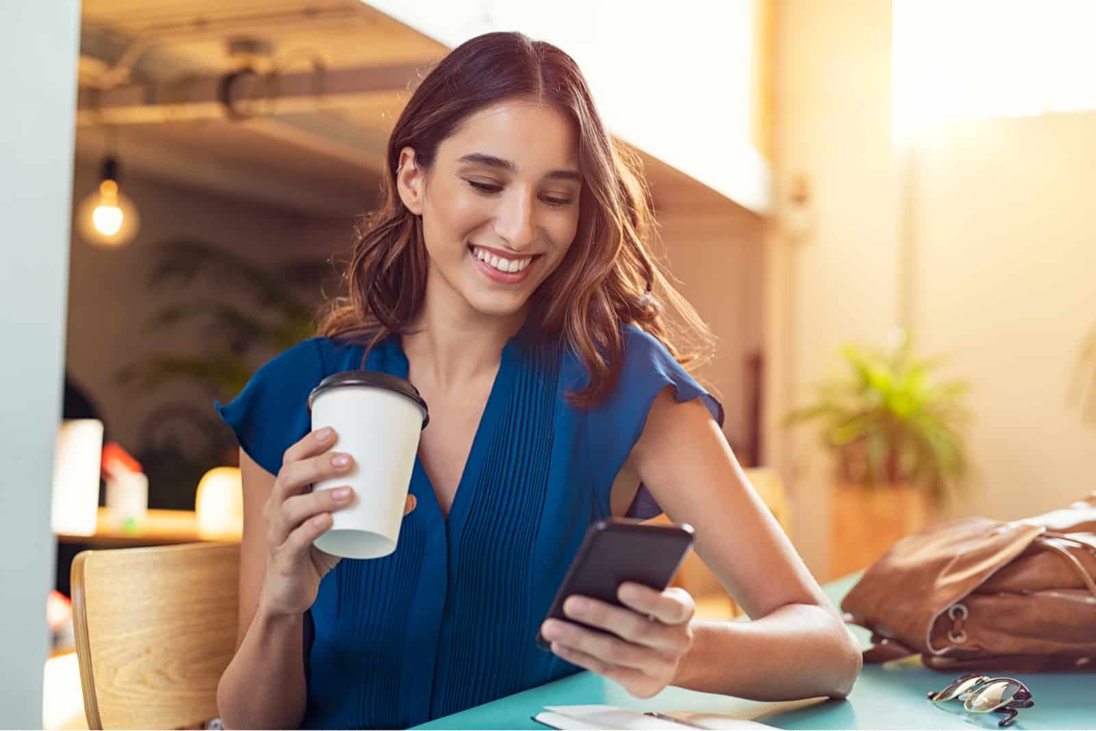 a young smiling woman sits at a table drinking coffee and holding a phone in her hand