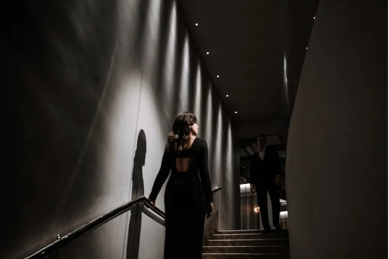 beautiful brunette rises from the stairs in black dress with a man in suit waiting at the top