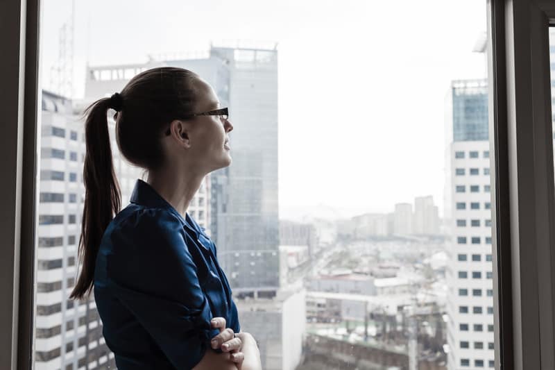 businesswoman standing near the glasswall of the tall building looking at the skies smiling