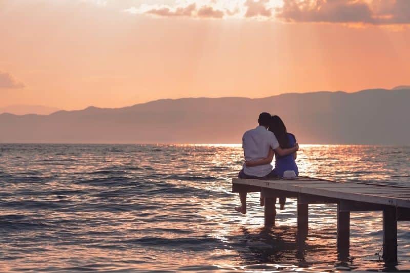 casal a apreciar o pôr do sol sentado numa plataforma de madeira sobre o mar