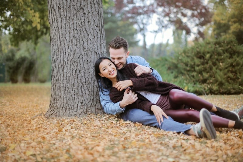 uomo e donna sorridenti seduti sulle foglie sotto l'albero