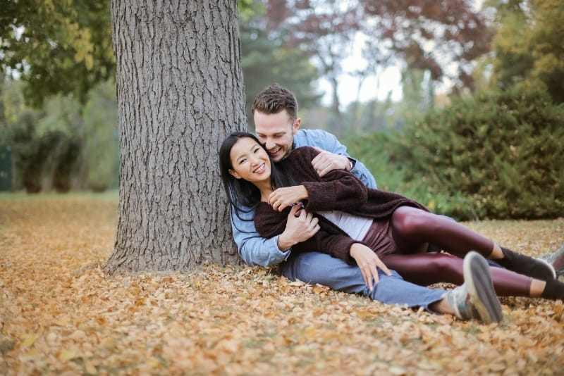 hombre y mujer sonrientes sentados sobre hojas bajo el árbol