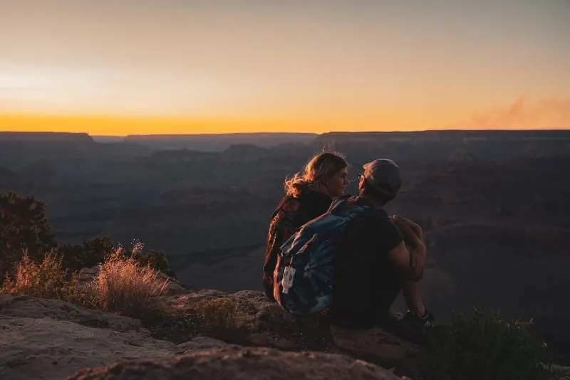 man with backpack and woman sitting on rock during sunset
