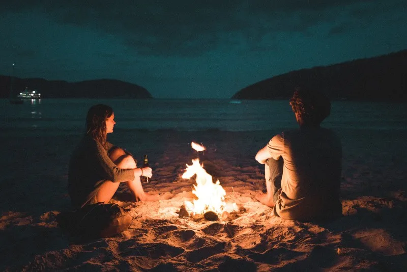 man and woman sitting on seashore near bonfire