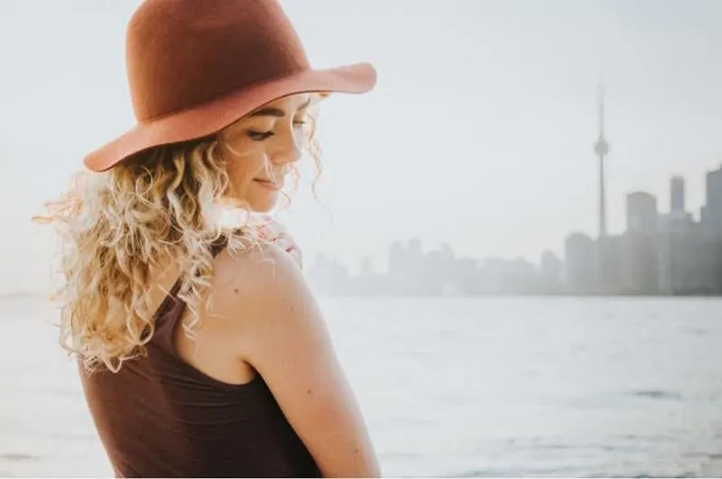 curly haired woman with a hat standing near a body of water facing the city