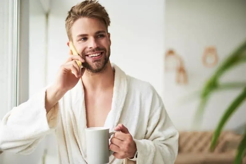 delighted young man talking on the mobile phone while holding a cup of coffee in the morning