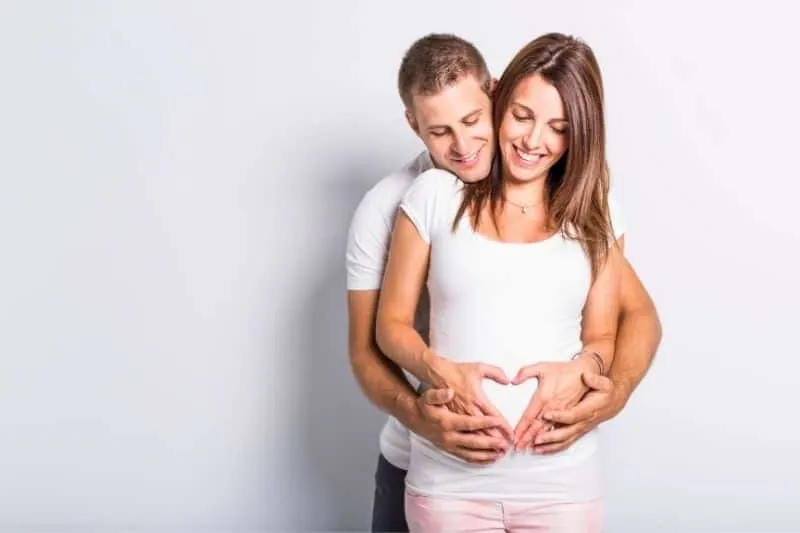 expecting baby couple hugging and standing against white wall