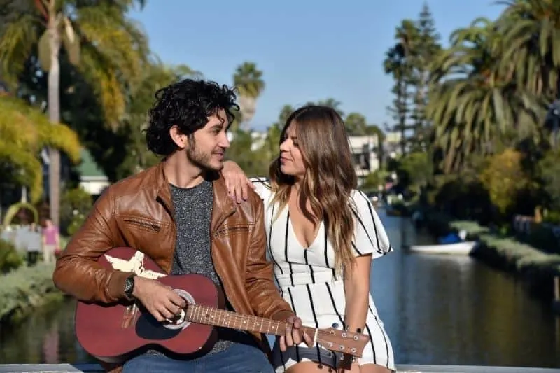 handsome man playing guitar with a woman smiling and sitting near the river bank