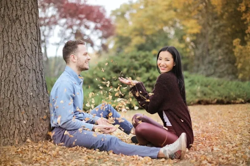 happy couple scattering dried leaves in the park while sitting on the ground