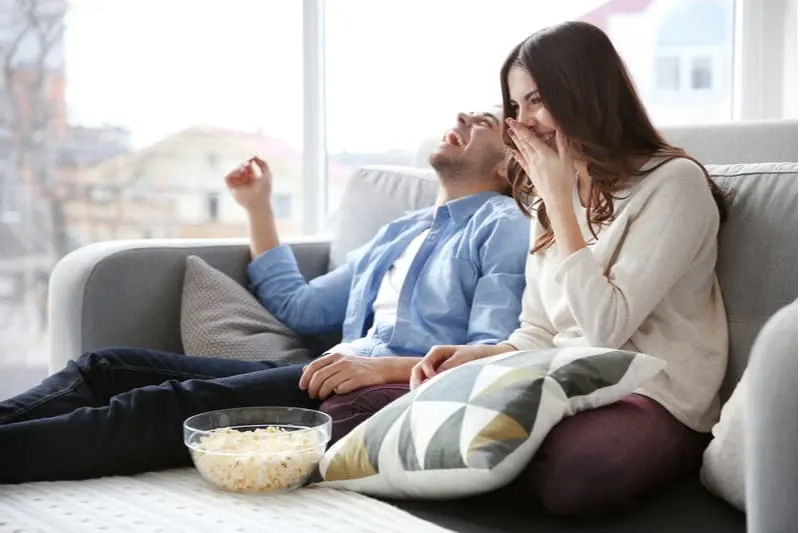 man and woman watching tv on the couch laughing and eating popcorn