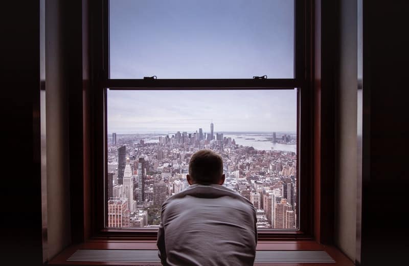 man in gray shirt looking at the city thru the window