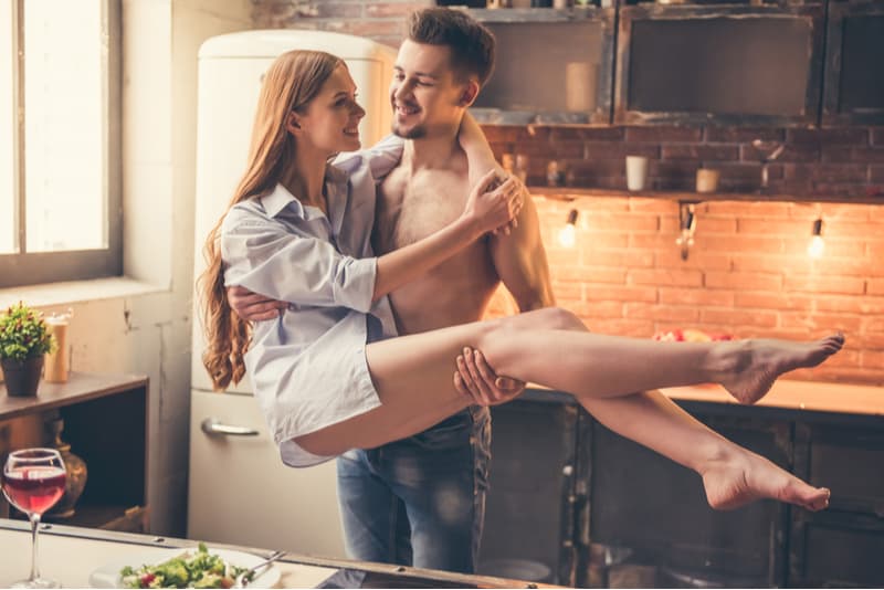 muscular half naked man carrying the woman wearing polo inside the kitchen