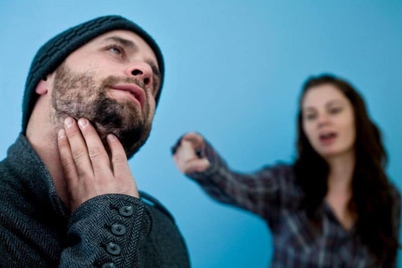 nagging woman pointing at a stressed man in focus standing near teal wall