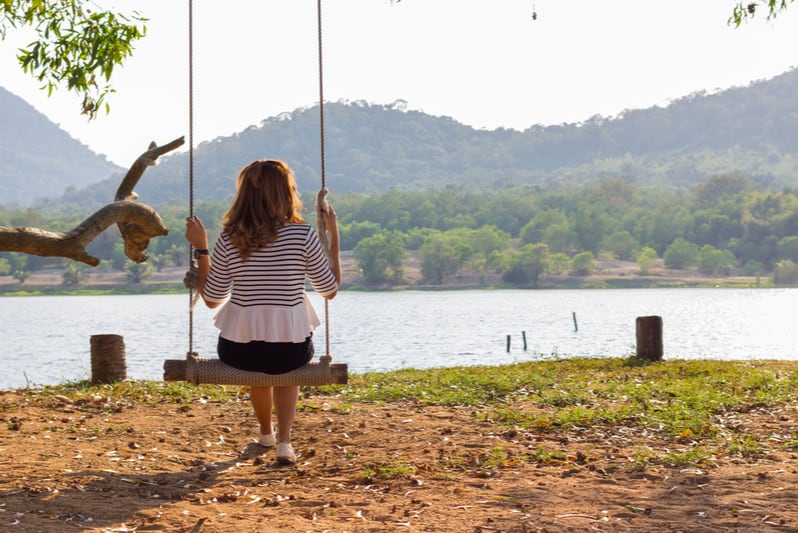 vista traseira de uma mulher triste sentada num baloiço perto de um corpo de água