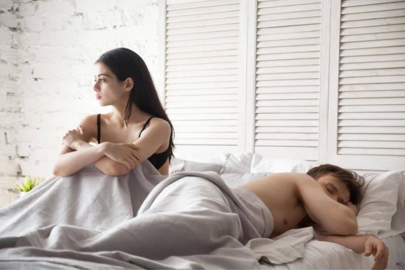 sad and frustrated woman wearing bras sitting beside a man sleeping half naked