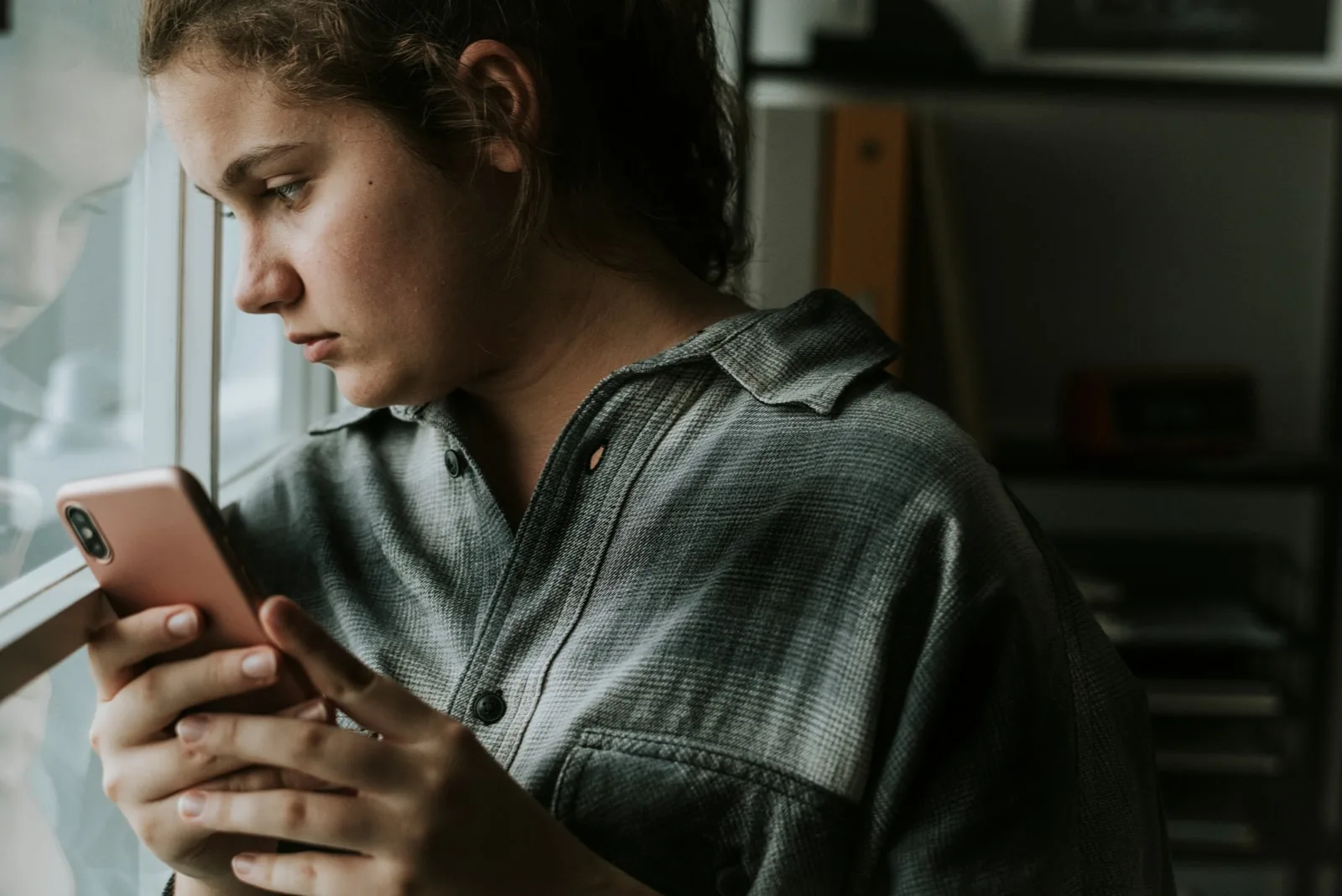sad girl with cell phone in hand standing by the window