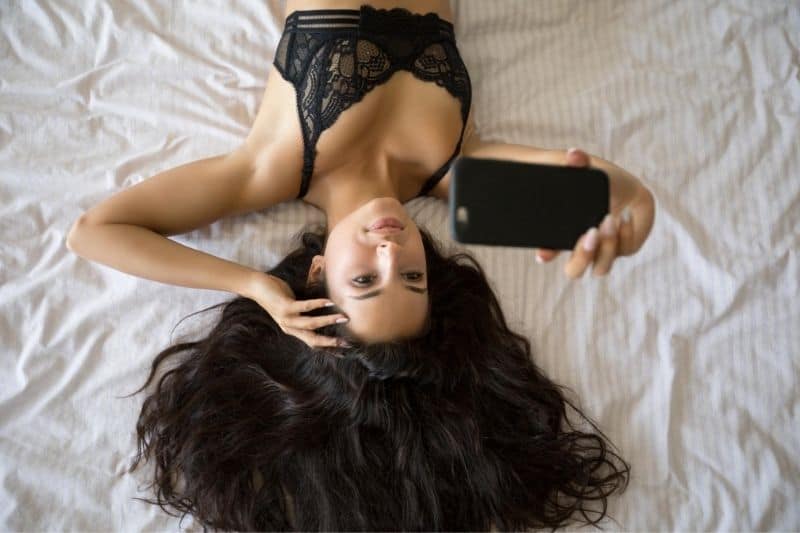 sexy woman lying down in bed wearing bras taking a selfie